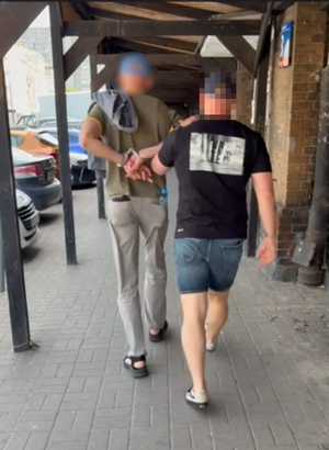 Zdjęcie przedstawia ubranego w ciemną koszulkę policjanta i krótkie niebieskie spodenki oraz czarną czapkę, który prowadzi po swojej lewej stronie zatrzymanego mężczyznę. Mężczyzna ten ma kajdanki założone na rękach trzymanych z tyłu. Obaj są tyłem do aparatu.