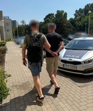 Zdjęcie przedstawia ubranego w cywilne ubrania policjanta, który ma na lewym przedramieniu odblaskową opaskę z napisem POLICJA. Policjant prowadzi po swojej prawej stronie. przez dziedziniec, zatrzymanego mężczyznę. Ich twarze są zamazane komputerowym filtrem. Obaj znajdują się tyłem do obiektywu aparatu. Zatrzymany mężczyzna ma kajdanki założone na rękach trzymanych z tyłu. Po ich prawej stronie widać zaparkowany radiowóz.