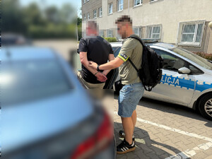 Zdjęcie przedstawia ubranego w cywilne ubrania policjanta, który ma na lewym przedramieniu odblaskową opaskę z napisem POLICJA. Policjant umieszcza w samochodzie zatrzymanego mężczyznę. Ich twarze są zamazane komputerowym filtrem. Obaj znajdują się lewym bokiem do obiektywu aparatu. Zatrzymany mężczyzna ma kajdanki założone na rękach trzymanych z tyłu. Po ich prawej stronie widać zaparkowany radiowóz.