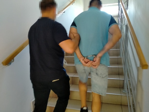 Zdjęcie przedstawia ubranego na ciemno policjanta, który wprowadza po schodach, po swojej prawej stronie zatrzymanego. Mężczyzna ten ma błękitną koszulkę na krótki rękaw i kajdanki na rękach trzymanych z tyłu. Ich twarze są zasłonięte komputerowym filtrem. Obaj znajdują się tyłem do obiektywu aparatu.