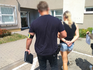 Zdjęcie przedstawia ubranego na ciemno policjanta, który prowadzi przez dziedziniec, po swojej prawej stronie zatrzymaną. Kobieta ma czarną bluzkę na ramiączkach i niebieskie krótkie spodenki jeansowe. Ma kajdanki na rękach trzymanych z tyłu. Ich twarze są zasłonięte komputerowym filtrem. Oboje znajdują się tyłem do obiektywu aparatu. Przed nimi widać budynek komendy i okratowane okno.
