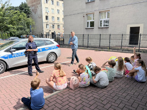 Zdjęcie przedstawia policjantkę i mężczyznę stojących przed dziećmi siedzącymi na chodniku, na dziedzińcu szkoły.