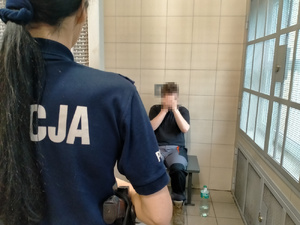 Zdjęcie przedstawia umundurowaną policjantkę, która stoi w otwartych drzwiach celi, tyłem do obiektywu aparatu. Zatrzymany mężczyzna siedzi na ławce, na wprost wejścia. Mężczyzna ten jest ubrany na ciemno.