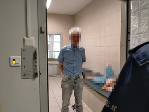 Zdjęcie przedstawia fragment sylwetki umundurowanego policjanta, stojącego w otwartych drzwiach celi. Przed nim stoi mężczyzna, którego twarz jest zablurowana komputerowym filtrem.