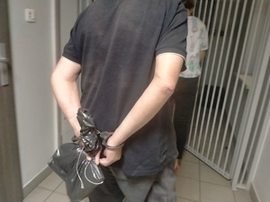 Zdjęcie przedstawia policjantkę ubraną w cywilne ubrania, która idzie przed zatrzymanym i otwiera drzwi do pomieszczenia dla osób zatrzymanych. Za nią idzie zatrzymany mężczyzna, ubrany na ciemno, który ma kajdanki założone na rękach trzymanych z tyłu. Mężczyzna trzyma w dłoniach ciemny foliowy woreczek.