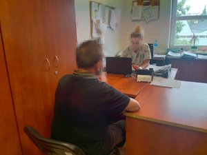 Zdjęcie przedstawia policjantkę ubraną w cywilne ubrania, która siedzi przed monitorem komputera. Na przeciwko niej, po drugiej stronie biurka siedzi zatrzymany mężczyzna. Ich twarze są zniekształcone komputerowym filtrem.