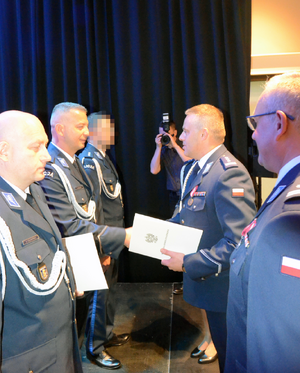 Zdjęcie przedstawia I Zastępcę Komendanta Stołecznego Policji wręczającego akt minowania policjantowi stojącemu w rzędzie z innymi policjantami.