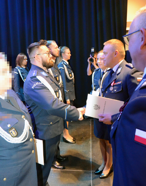 Zdjęcie przedstawia I Zastępcę Komendanta Stołecznego Policji wręczającego akt minowania policjantowi stojącemu w rzędzie z innymi policjantami.