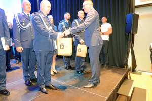 Zdjęcie przedstawia mężczyznę w garniturze, który wręcza torebki z upominkami dwóm policjantom.