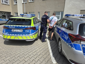 Zdjęcie przedstawia policjanta prowadzącego zatrzymanego mężczyznę pomiędzy dwoma zaparkowanymi radiowozami.
