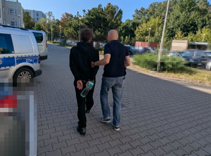 Zdjęcie przedstawia policjanta ubranego w ciemną koszulkę na krótki rękaw i spodnie jeansowe, który prowadzi po swojej lewej stronie zatrzymanego mężczyznę. Młody mężczyzna jest ubrany na ciemno. Na rękach trzymanych z tyłu ma założone kajdanki. Obaj mają zasłonięte głowy komputerowym filtrem. Wokół widać zaparkowane pojazdy.