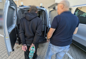 Zdjęcie przedstawia policjanta ubranego w ciemną koszulkę na krótki rękaw i spodnie jeansowe, który stoi przed otwartymi tylnymi drzwiami radiowozu z zatrzymanym mężczyzną. Młody mężczyzna jest ubrany na ciemno. Na rękach trzymanych z tyłu ma założone kajdanki. Obaj mają zasłonięte głowy komputerowym filtrem. Wokół widać zaparkowane pojazdy.