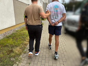 Zdjęcie przedstawia ubranego w brązową koszulkę na krótki rękaw policjanta, który prowadzi po swojej prawej stronie, po chodniku zatrzymanego mężczyznę. Ich twarze są zasłonięte komputerowym filtrem.