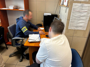 Zdjęcie przedstawia policjanta ubranego w odblaskową opaskę z napisem POLICJA, na prawym przedramieniu, który siedzi w pomieszczeniu służbowym, przy biurku, przed monitorem komputera. Zatrzymany mężczyzna siedzi przy biurku tyłem do obiektywu aparatu. Jest ubrany w białą koszulkę na krótki rękaw.