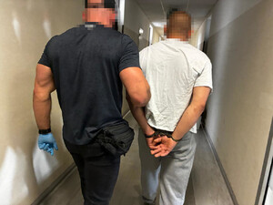 Zdjęcie przedstawia ubranego na ciemno policjanta, w niebieskich ochronnych rękawiczkach, który prowadzi po korytarzu, po swojej prawej stronie zatrzymanego mężczyznę, ubranego w białą koszulkę na krótki rękaw. Mężczyzna ten ma kajdanki założone na rękach trzymanych z tyłu.