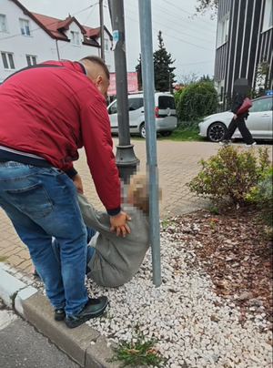 Zdjęcie przedstawia ubranego w czerwoną kurtkę i niebieskie spodnie jeansowe policjanta pochylającego się nad kobietą siedzącą na ziemi.