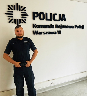 Zdjęcie przedstawia umundurowanego policjanta stojącego pod logiem Policji i napisem na ścianie Komenda Rejonowa Policji Warszawa VI.