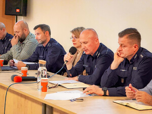 Zdjęcie przedstawia policjanta mówiącego do mikrofonu, który siedzi przy długim stole z innymi policjantami oraz osobami spoza Policji. Naprzeciwko nich siedzą osoby na krzesłach.
