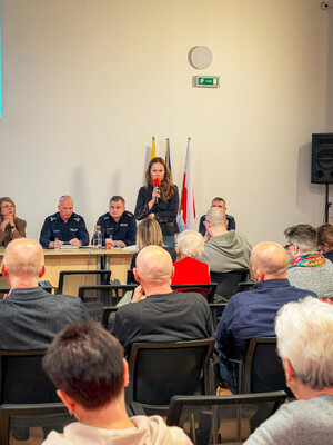 Zdjęcie przedstawia kobietę mówiącą do mikrofonu, która stoi przy policjantach siedzących przy długim stole. Naprzeciwko nich siedzą osoby na krzesłach.