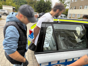Zdjęcie przedstawia policjanta ubranego w ciemny bezrękawnik i niebieską bluzę, który umieszcza na tylnym siedzeniu radiowozu  mężczyznę ubranego w jasną koszulkę, który ma kajdanki założone na rękach trzymanych z tyłu. Ich twarze są zasłonięte komputerowym filtrem. Po prawej stronie policjanta, stoi policjantka w ubraniu cywilnym.