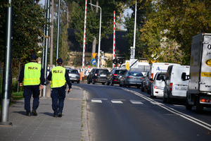 Zdjęcie przedstawia dwóch policjantów idących chodnikiem przy ulicy, na której stoją w ciągu samochody przed zamkniętym przejazdem kolejowym.