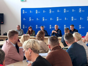 Zdjęcie przedstawia policjantów i kobietę siedzących za stołem naprzeciwko innych osób uczestniczących w spotkaniu.