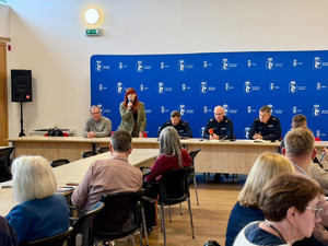 Zdjęcie przedstawia policjantów siedzących za stołem i stojącą z mikrofonem kobietę, naprzeciwko innych osób uczestniczących w spotkaniu.